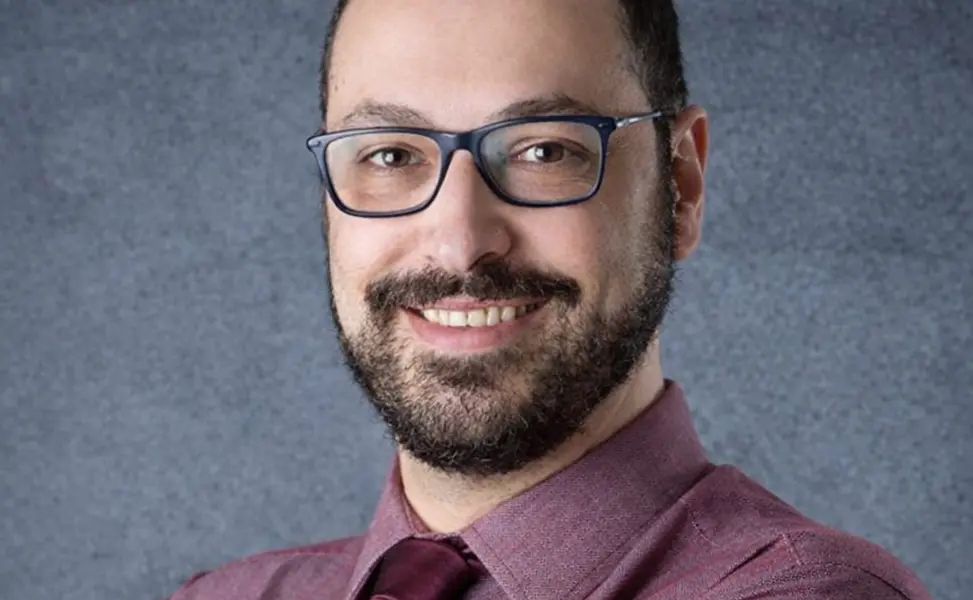Homem de meia-idade com barba, óculos de armação azul e camisa vinho, sorrindo levemente em fundo cinza neutro