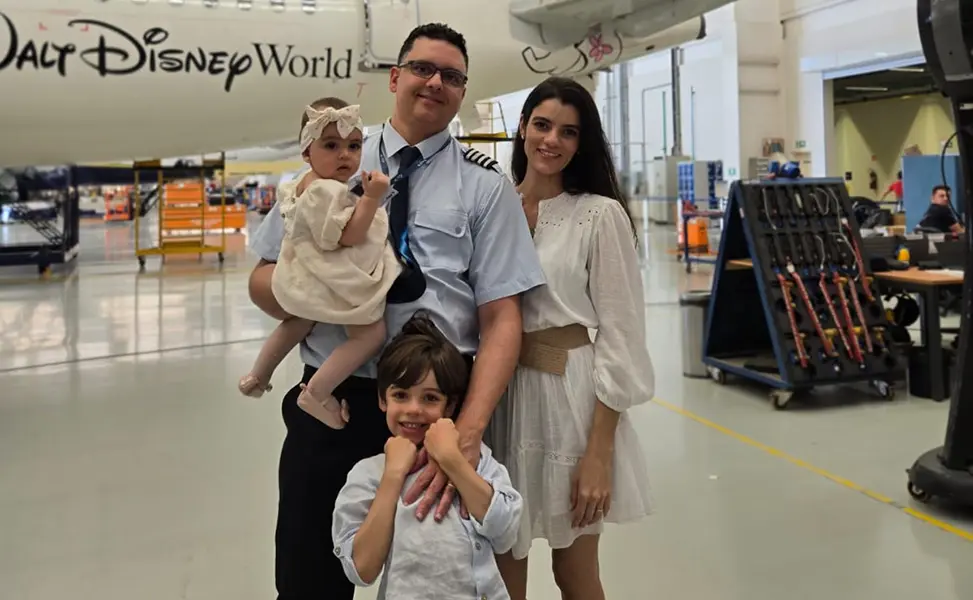 Homem com uniforme de piloto segurando bebê com laço na cabeça, mulher ao lado com vestido branco e menino à frente, posando em hangar com avião ao fundo com a inscrição Walt Disney World