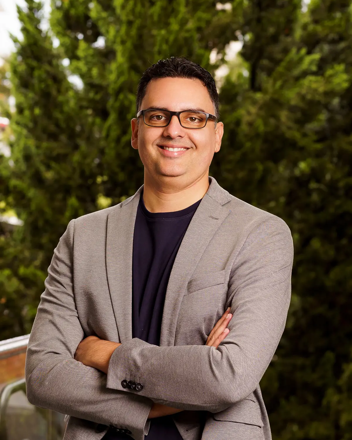 Homem de blazer cinza e camiseta preta, óculos de armação escura, braços cruzados e sorrindo, em ambiente externo com árvores verdes ao fundo