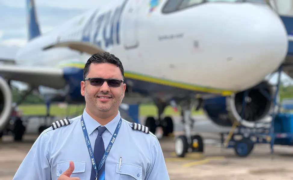 Homem com uniforme de primeiro-oficial da Azul, óculos de sol, lanyard da companhia, fazendo gesto de positivo com o polegar, em frente a aeronave da Azul em pátio externo