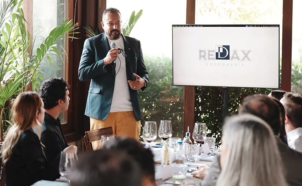 Felipe Xavier discursa para convidados em almoço de negócios, com a marca Redax Engenharia projetada ao fundo