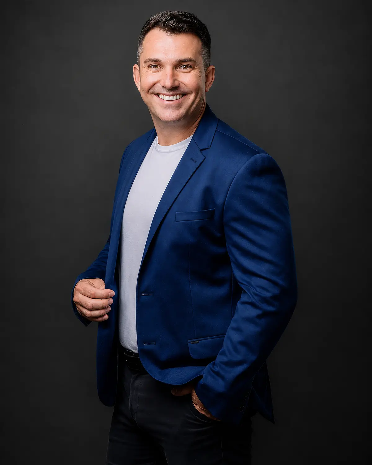 Homem sorrindo, usando blazer azul-marinho sobre camiseta branca, posando em estúdio com fundo cinza escuro