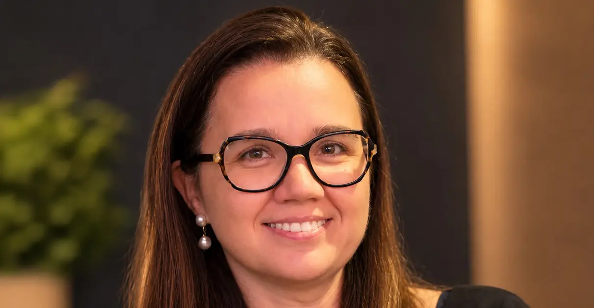 Mulher de cabelos castanhos, óculos de armação escura e brincos de pérola, sorrindo, com fundo desfocado em tons escuros e planta ao fundo