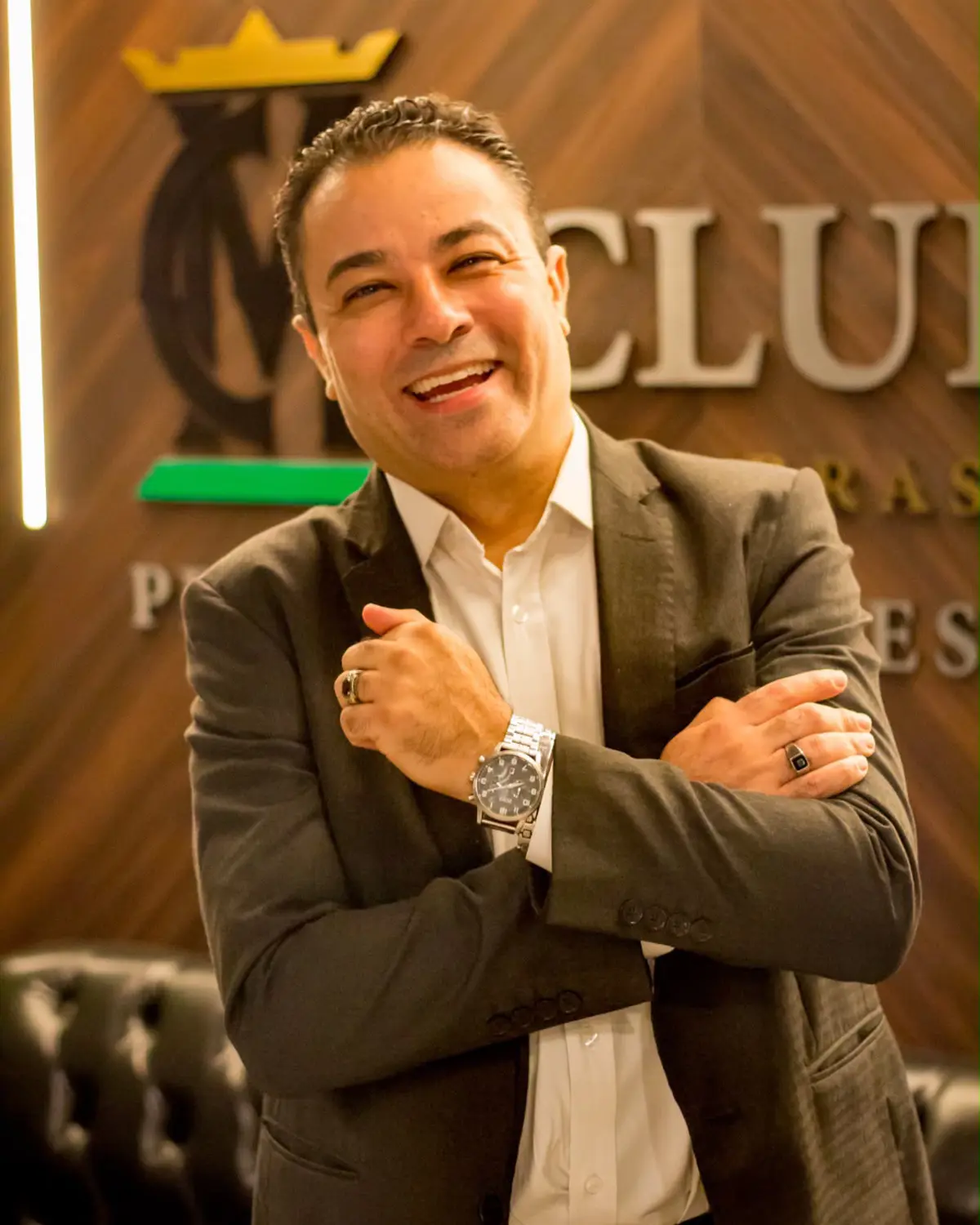 Homem sorrindo de braços cruzados, vestindo terno escuro e relógio prateado, em frente a painel com logotipo corporativo