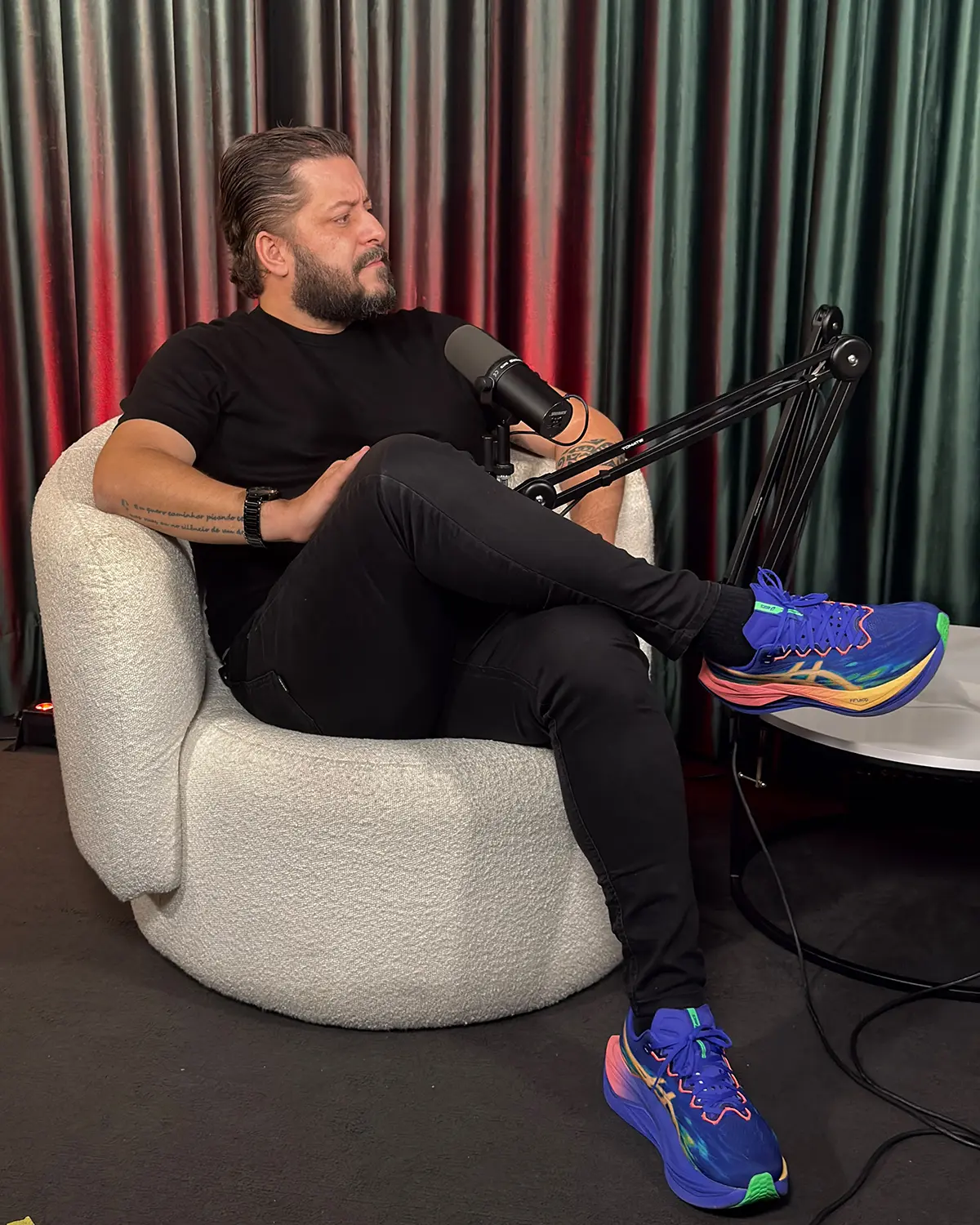 Homem de camiseta e calça pretas sentado em poltrona redonda clara, com microfone de podcast e tênis azul colorido