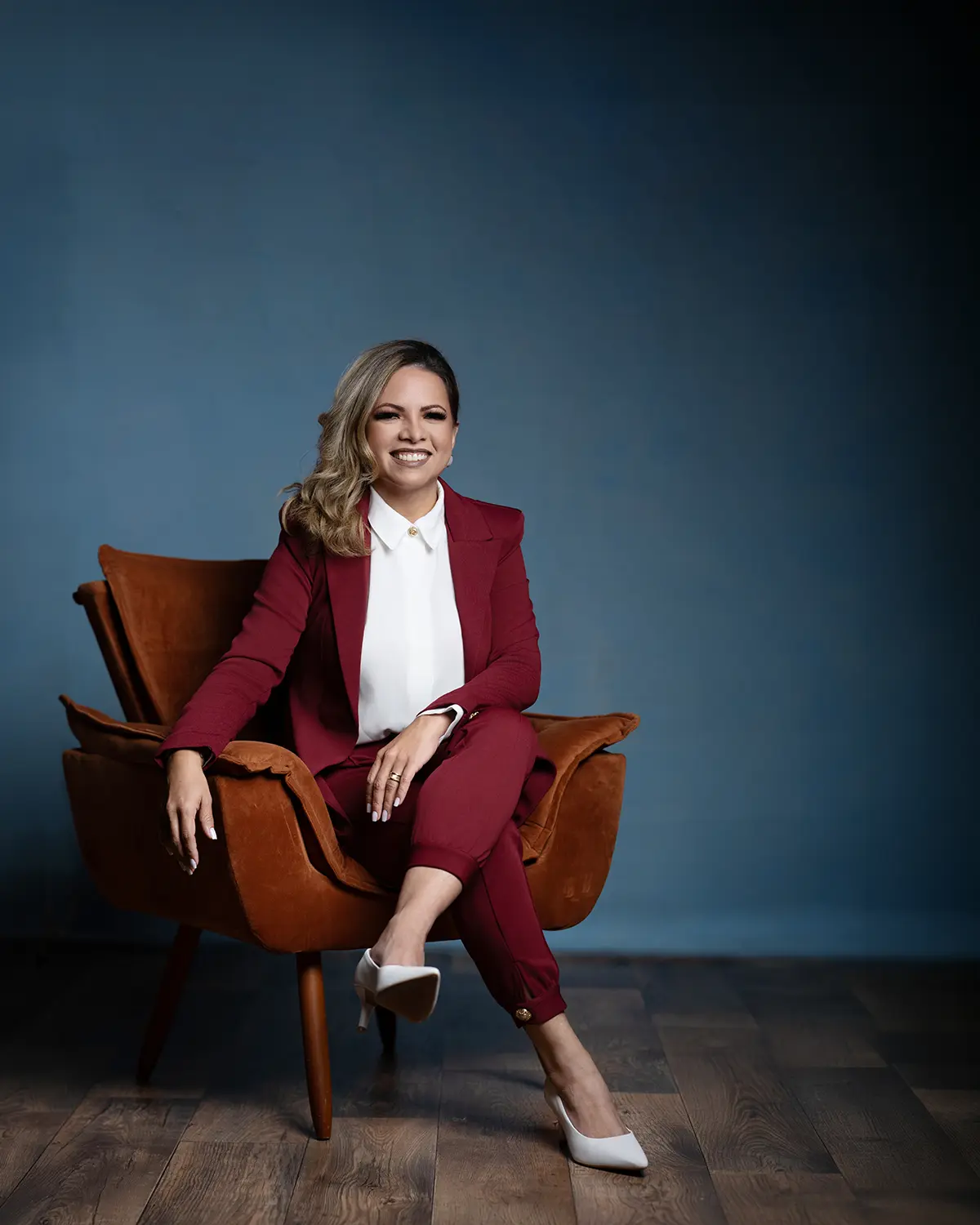 Woman in a burgundy suit and white blouse, seated in a caramel armchair and smiling in a dark-blue studio