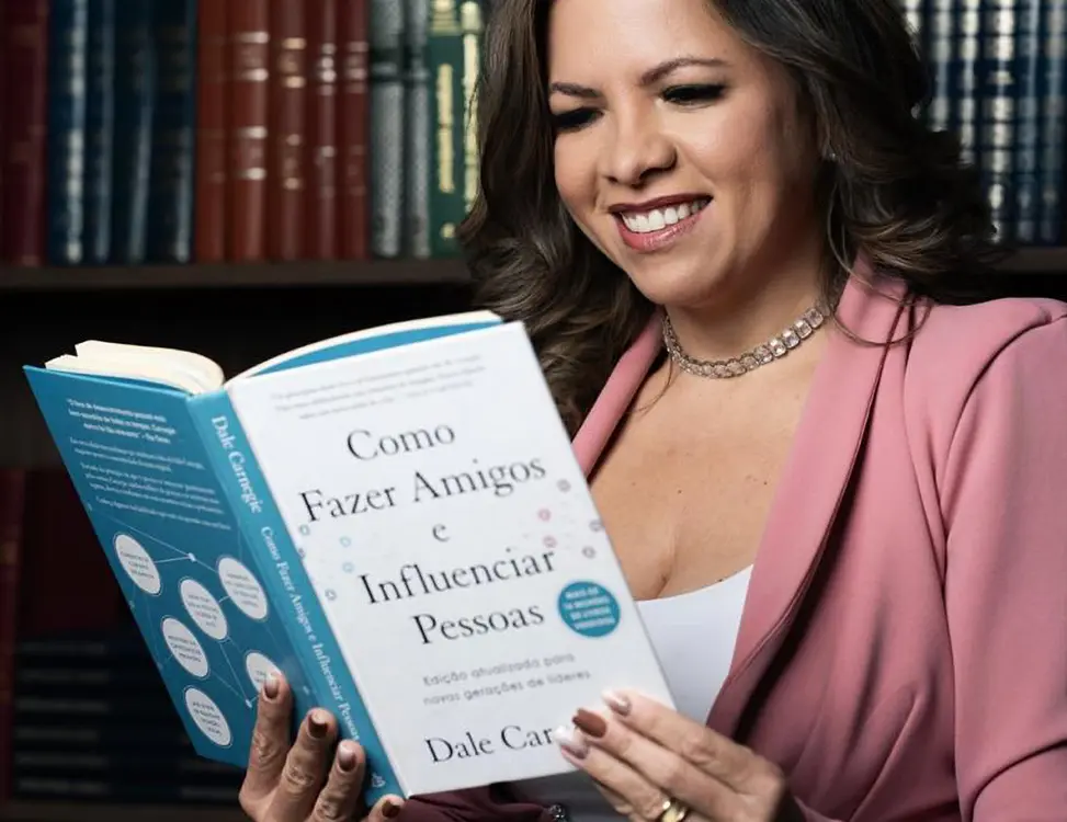 Taís Araújo smiles while leafing through Dale Carnegie’s book How to Win Friends and Influence People, with a bookcase in the background