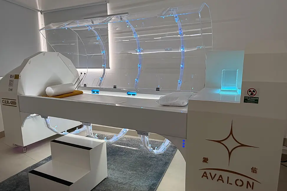 Equipamento médico branco modelo CER-GSD da marca Avalon com estrutura translúcida iluminada em azul e maca acolchoada, em sala com paredes brancas