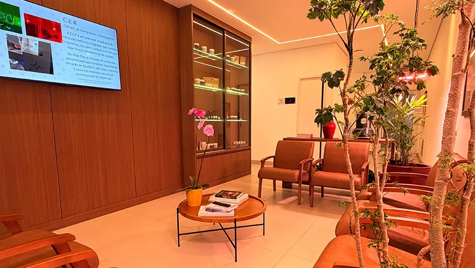 Recepção da Vitae Flux com poltronas de couro marrom, mesa de centro redonda com livros, armário iluminado com produtos, plantas e televisor na parede